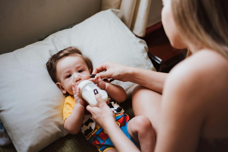 Mother feeding a baby with a bottle while sitting on a bed at home